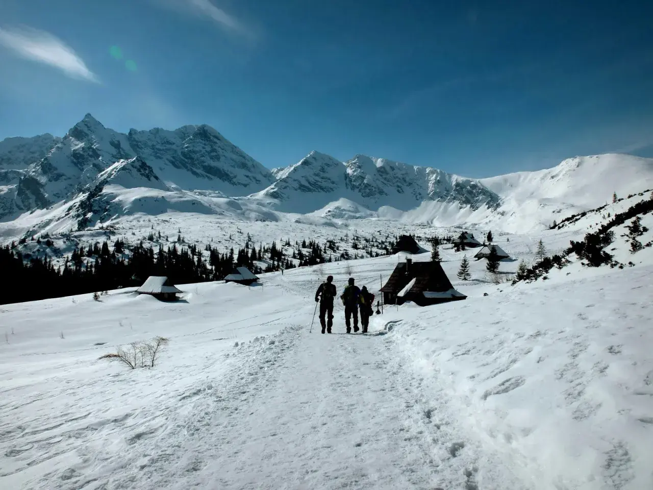 Trzej turyści idą szlakiem przez zaśnieżone Tatry zimą dla średniozaawansowanych, mijając góralskie chatki.
