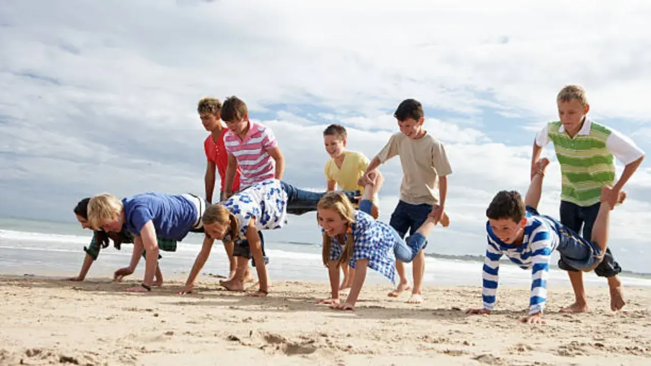 Niños compiten en juegos en la playa sin material, haciendo carreras de carretilla humana y otros desafíos divertidos.