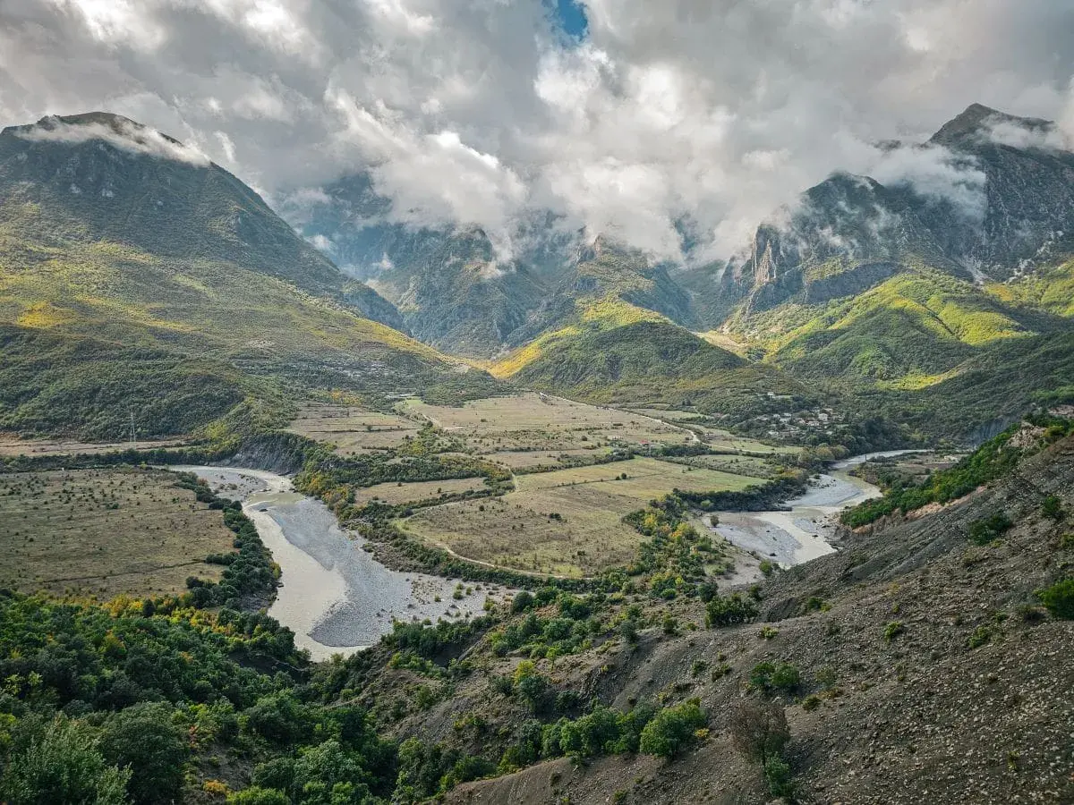 Ein malerischer Fluss schlängelt sich durch ein grünes Tal in Albanien, umgeben von bewaldeten Bergen unter dramatischen Wolken.