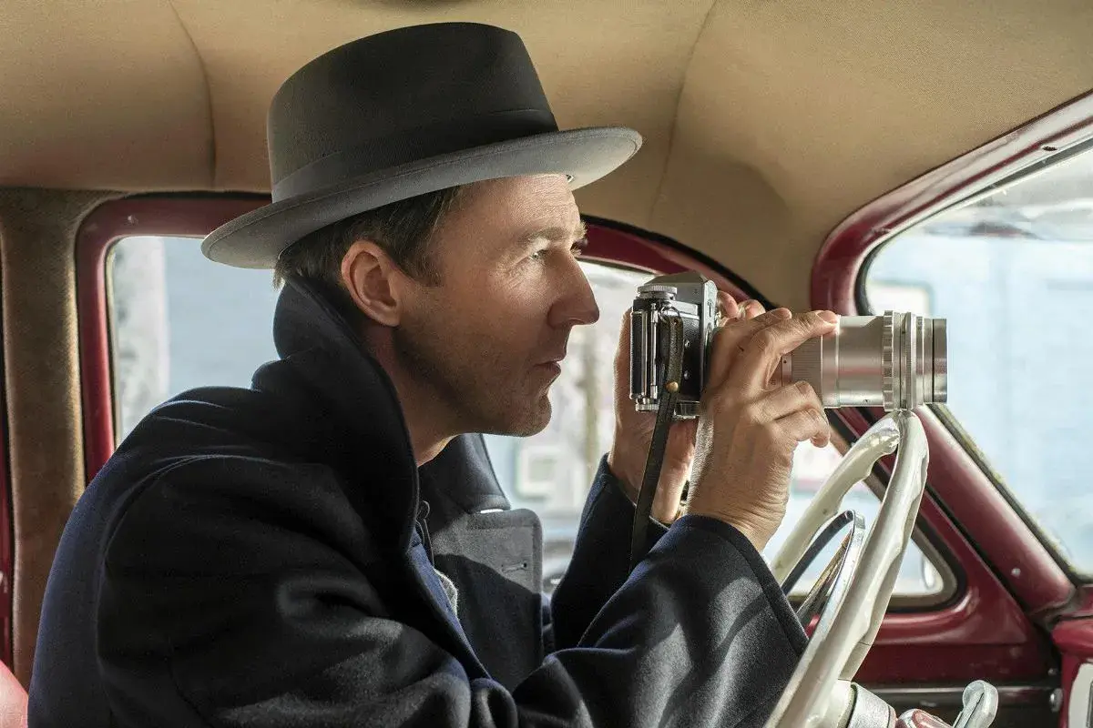 Edward Norton w filmach, w kapeluszu i płaszczu, siedzi za kierownicą samochodu i robi zdjęcie starym aparatem.