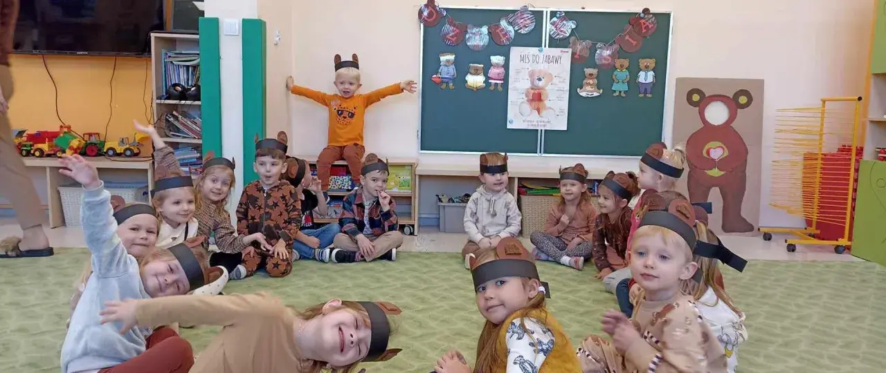 Dzieci w strojach misiów siedzą na dywanie w sali przedszkolnej.