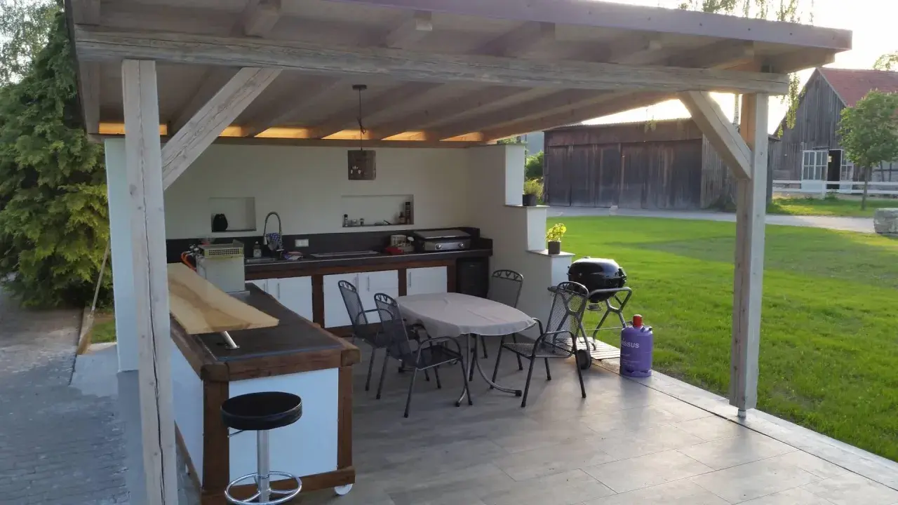 Gemütlicher **Grillplatz im Garten** mit Außenküche, Tisch und Stühlen unter einem Holzüberdach. Perfekt für Sommerabende.