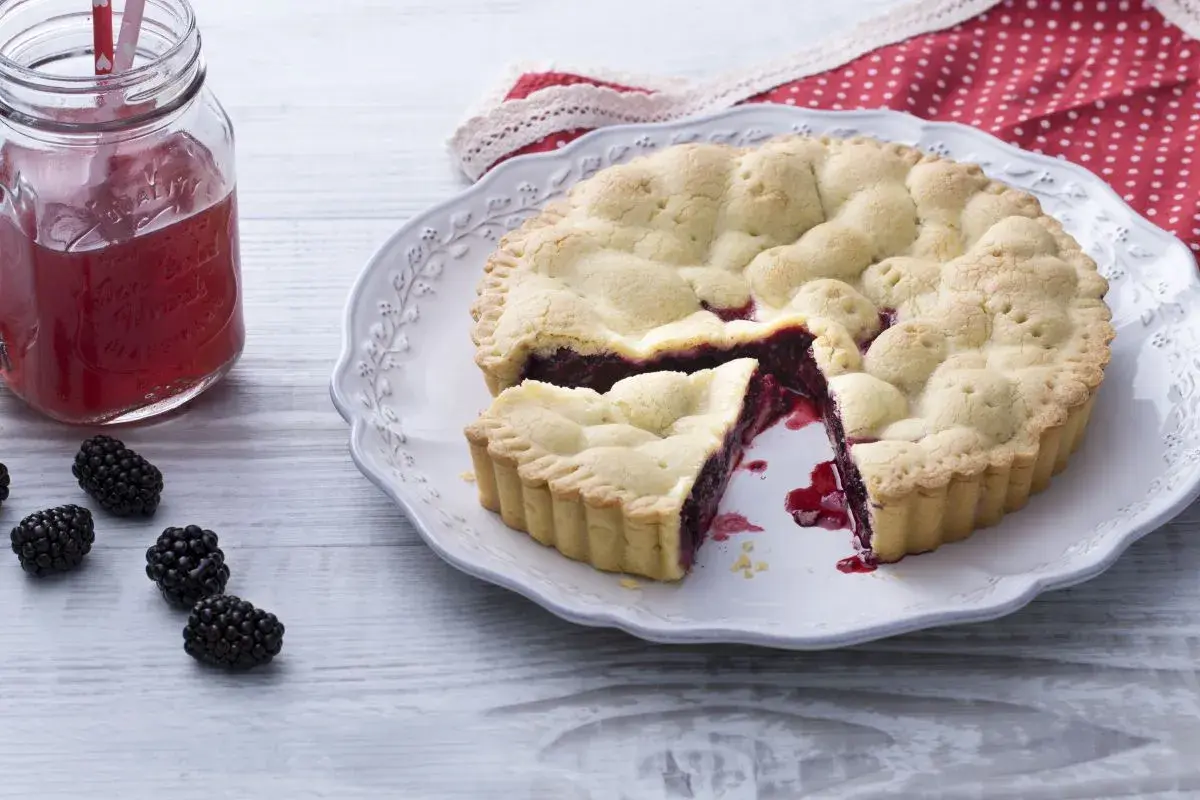 Une délicieuse tarte aux mûres, avec une part coupée, sur une assiette ornée. Des mûres fraîches et un verre de jus rouge complètent la scène.