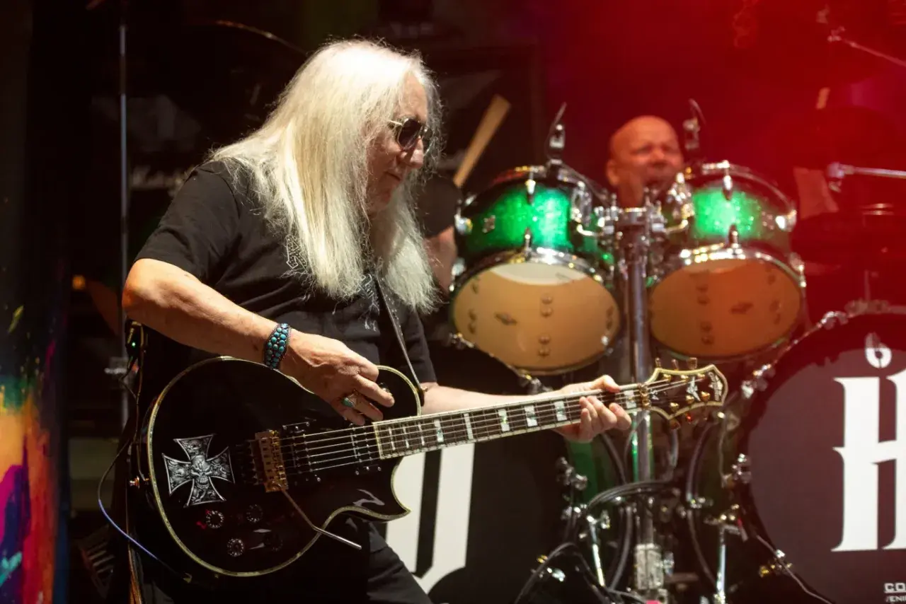 Uriah Heep członkowie na scenie. Gitarzysta z długimi blond włosami gra na czarnej gitarze z symbolem krzyża. W tle perkusista i zielone bębny.