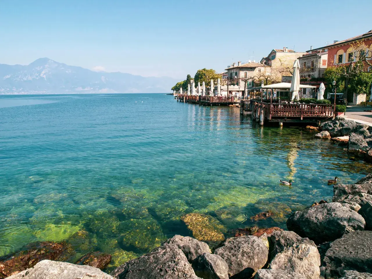 Lungolago di Gardone Riviera: cosa vedere? Case colorate, ristoranti con tavoli all'aperto e il lago cristallino.