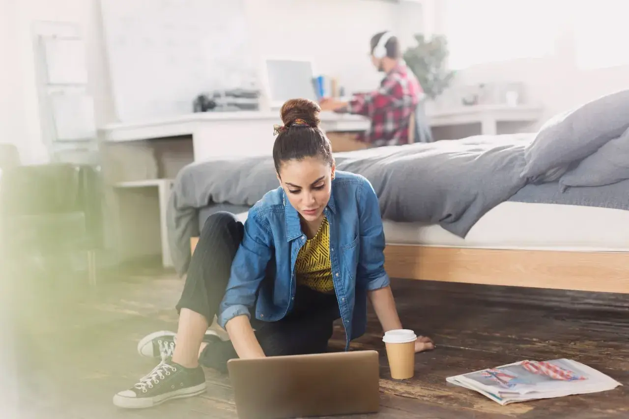 Młoda kobieta pracuje na laptopie na podłodze w pokoju. Obok niej kubek kawy i gazeta. W tle mężczyzna w słuchawkach. Zastanawia się, ile osób może mieszkać w wynajmowanym mieszkaniu.
