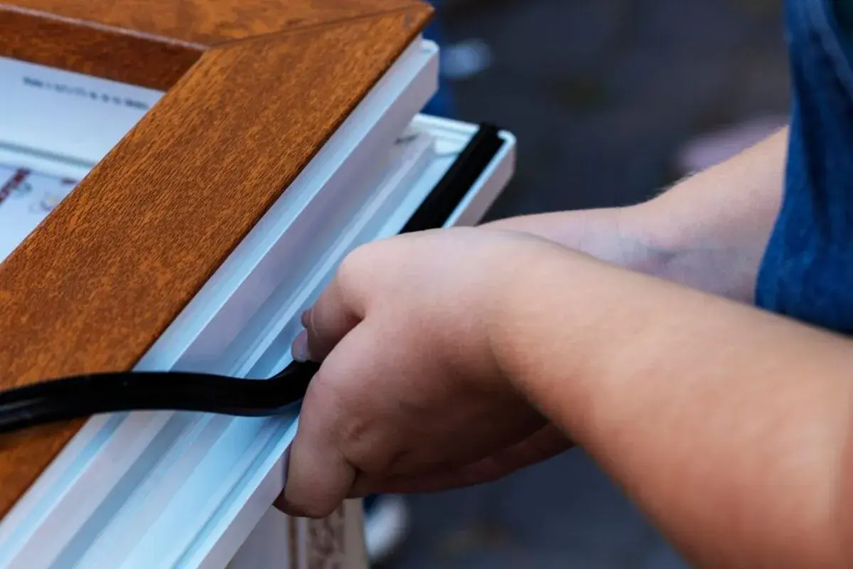 Pose d'un joint d'étanchéité pour comment insonoriser une porte. Les mains insèrent un joint noir dans le cadre blanc d'une porte imitation bois.