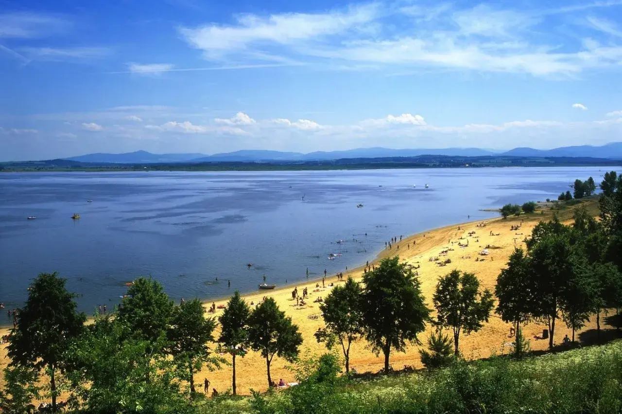 Słoneczna plaża nad Jeziorem Nyskim, pełna ludzi odpoczywających i kąpiących się. W oddali góry, a na wodzie żaglówki i łodzie. To idealne miejsce na wakacyjne atrakcje.