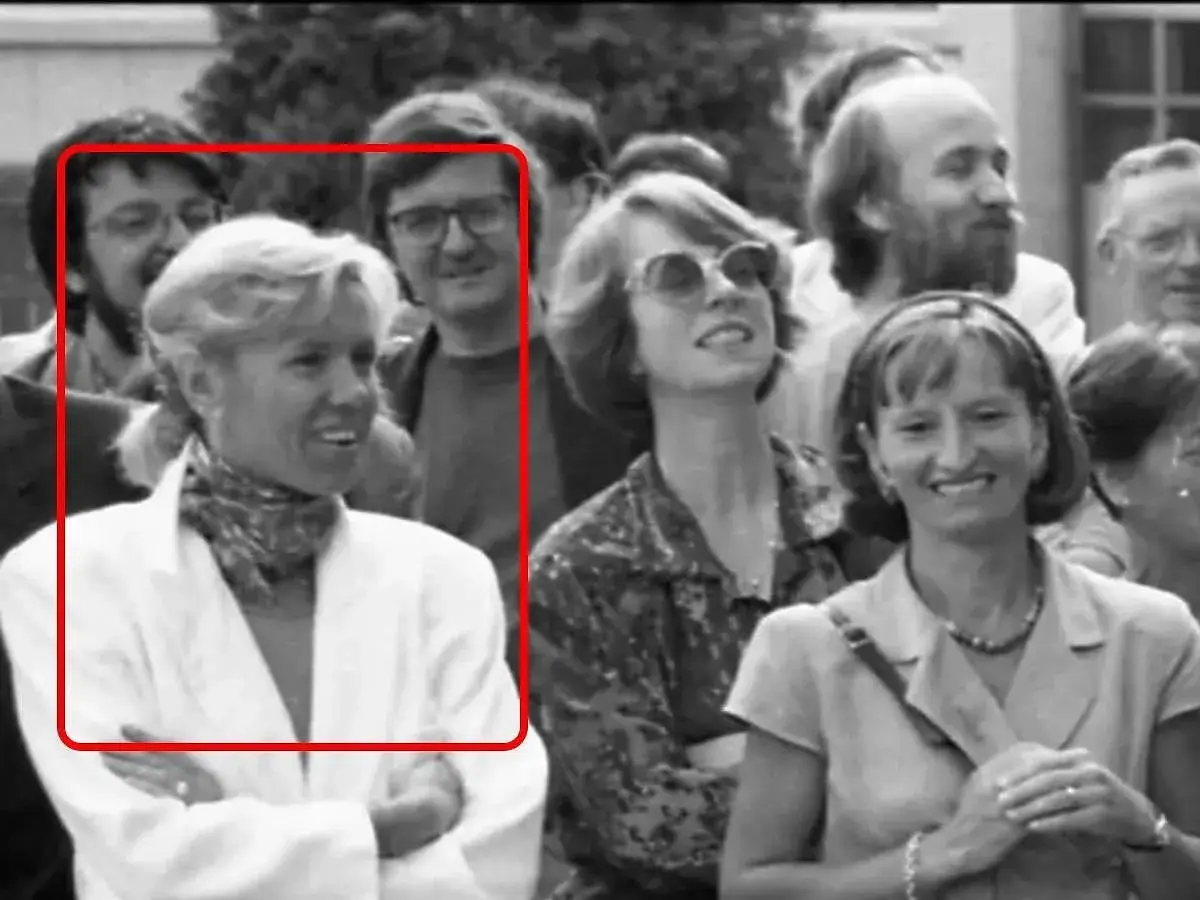 Brigitte Macron młoda, w białej marynarce i z apaszką na szyi, uśmiecha się do aparatu. W tle widać innych ludzi.
