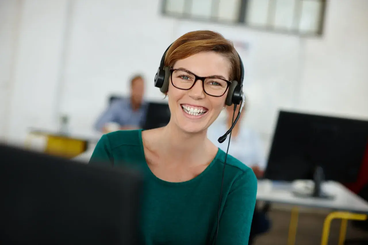 Uśmiechnięta kobieta w okularach i zestawie słuchawkowym, korzystająca z technologii VoIP w biurze.