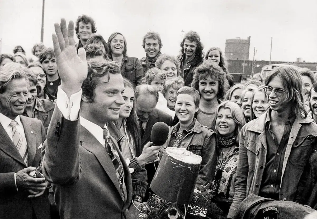 Kung Carl XVI Gustaf vinkar glatt till en folkmassa. En ung kvinna håller fram en mikrofon, kanske för att intervjua en av kungens barn eller någon annan i den glada gruppen.