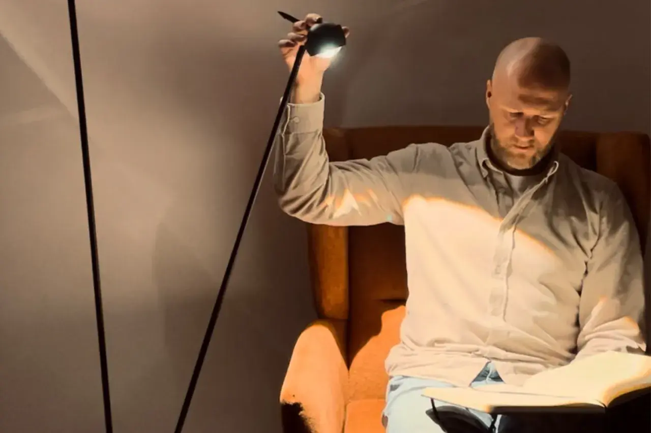 Hombre calvo sentado en sillón leyendo un libro, iluminado por un flexo de pie.