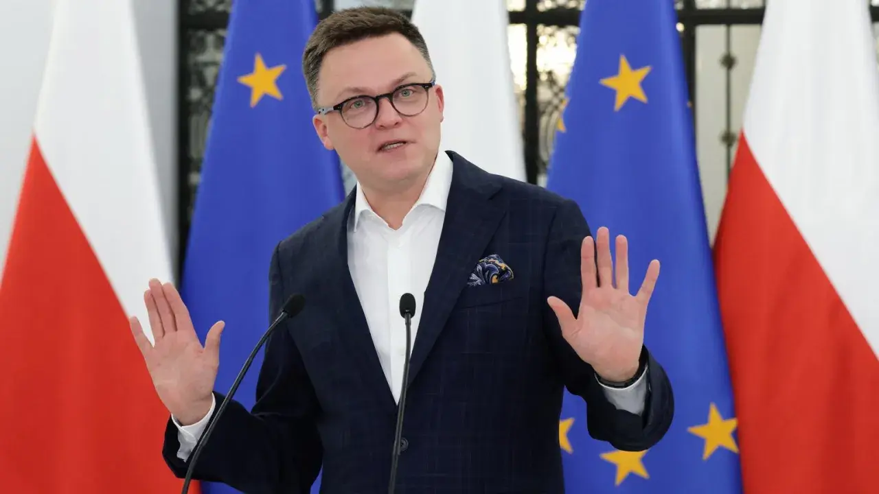 Szymon Hołownia w garniturze przemawia przed flagami Polski i UE.
