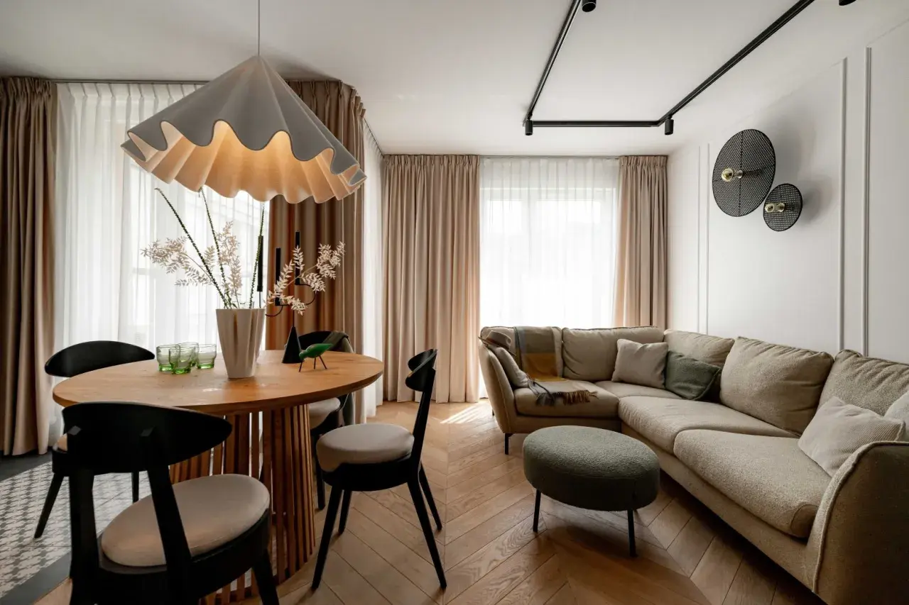 Przytulny salon z beżową kanapą i okrągłym stołem. Nad kanapą w salonie wiszą dekoracyjne lampy, a obok stoi zielona pufa.