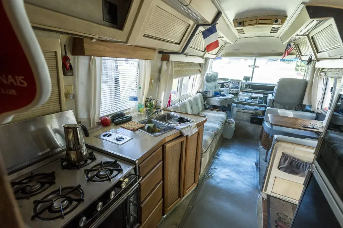 Vue de l'intérieur d'un camping-car américain : cuisine avec cuisinière, évier, et banquette. Drapeaux français et américain visibles.