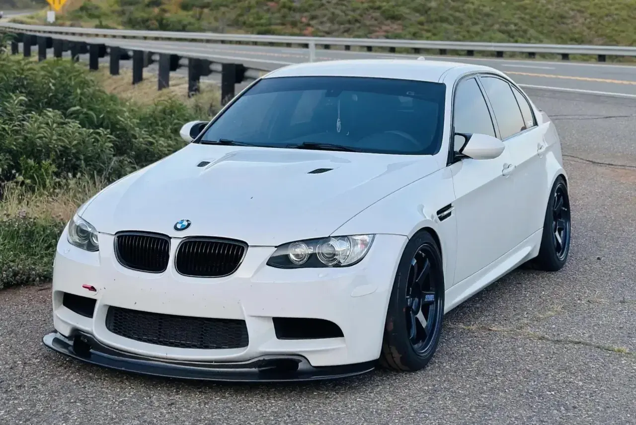 Białe BMW E90 M3 z czarnymi felgami i spoilerem, zaparkowane przy drodze z barierkami.