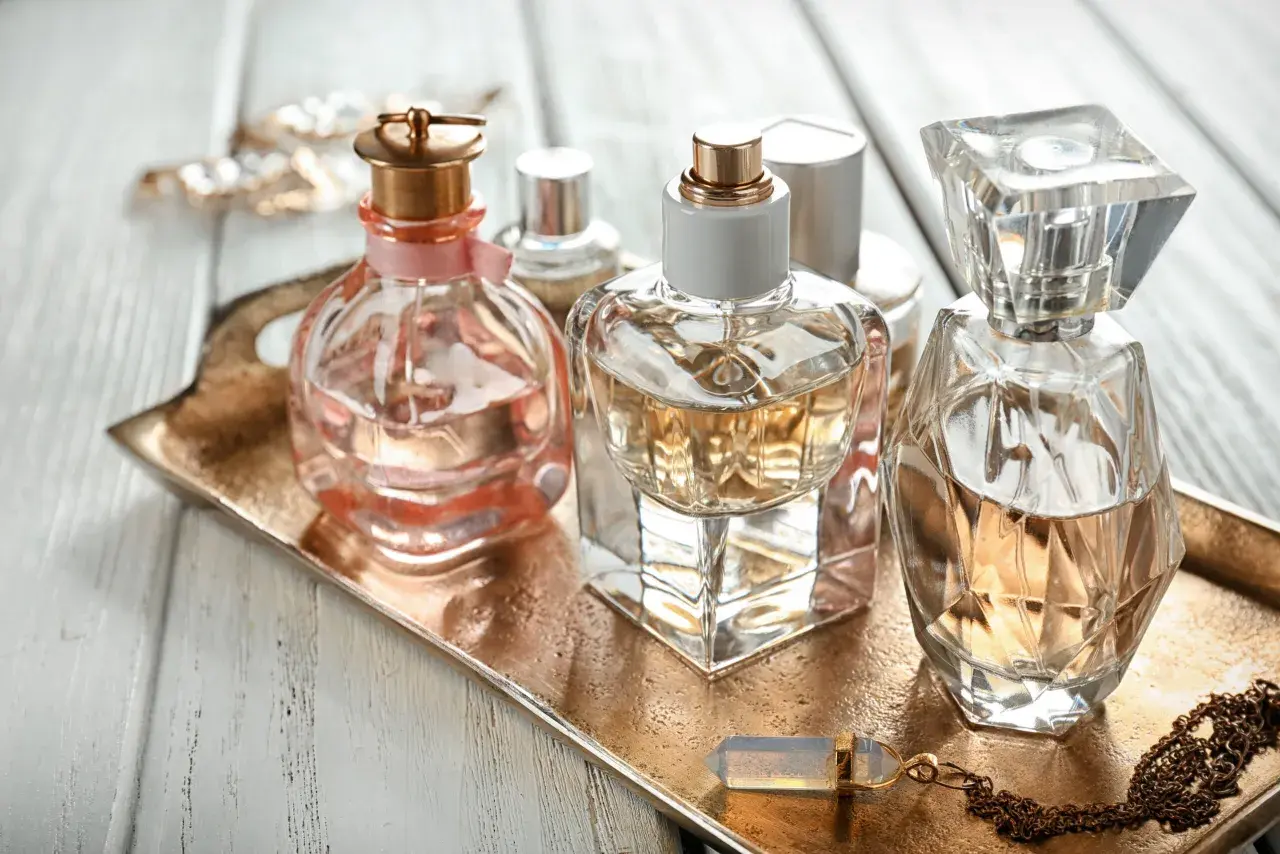 Eleganckie flakoniki perfum, w tym jeden z delikatną różową wstążką, stoją na złotej tacy. W tle błyszcząca biżuteria. Zapach, być może z nutą hexyl cinnamal, unosi się w powietrzu.