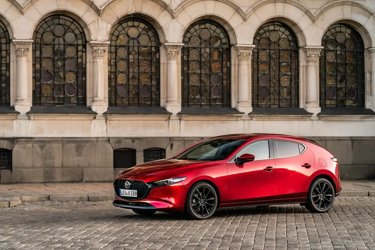 Un elegante Mazda 3 Skyactiv X rojo, con sus líneas fluidas, se detiene frente a una fachada histórica con arcos y vidrieras.