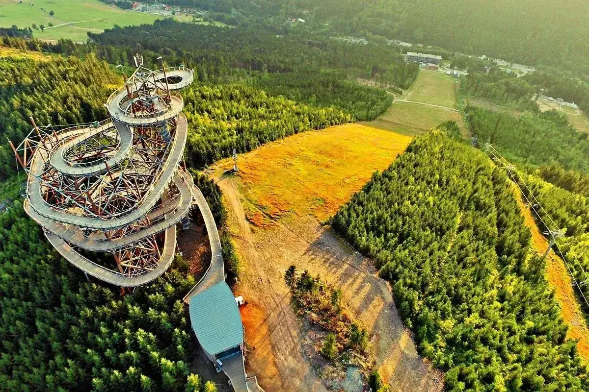 Widok z lotu ptaka na spiralny, drewniany wiszący most w Czechach, otoczony zielonym lasem i złotym polem.