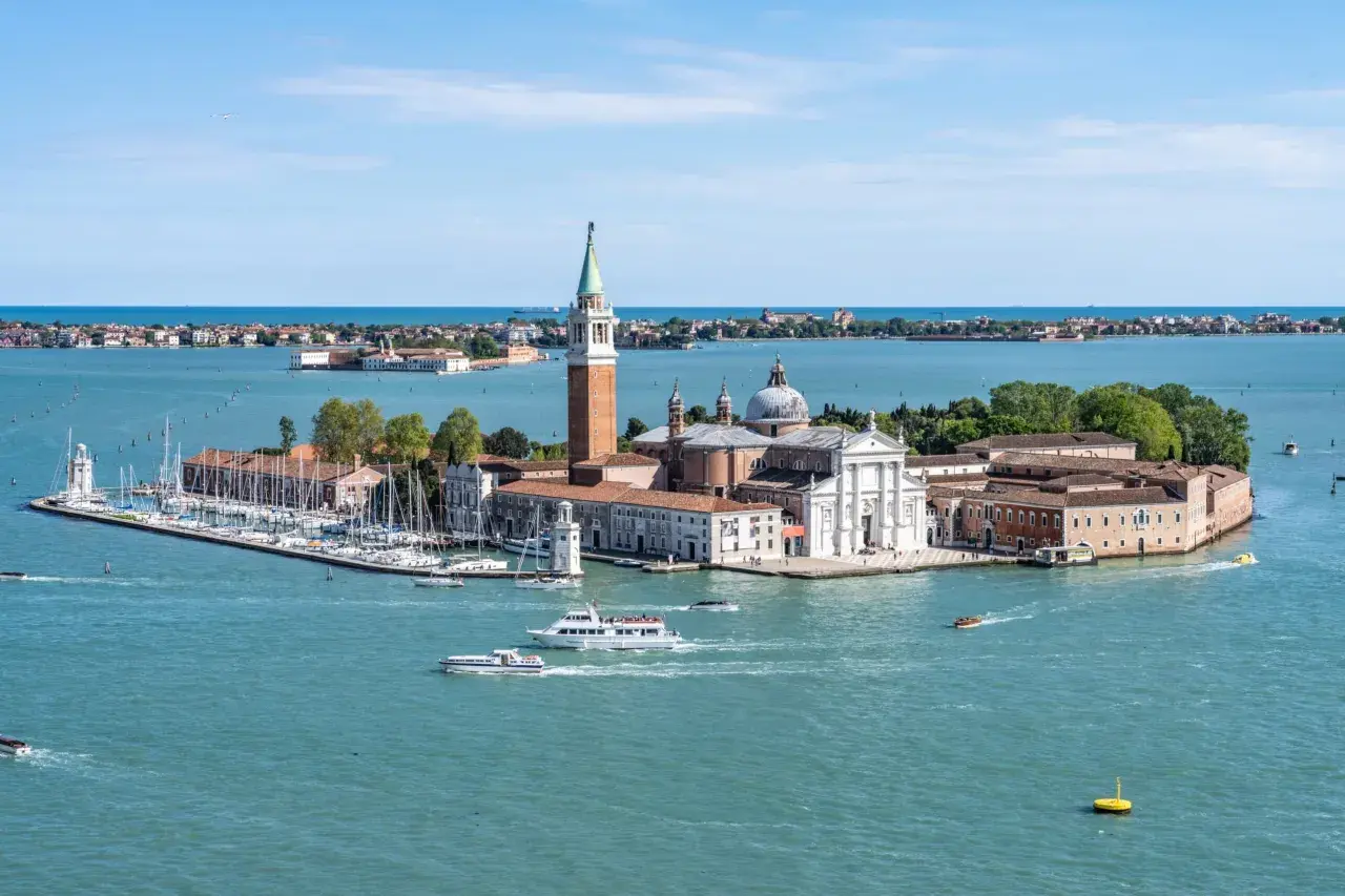 Widok na wyspę San Giorgio Maggiore w Wenecji, z kościołem i dzwonnicą. Wokół pływają łodzie, w tym prom.