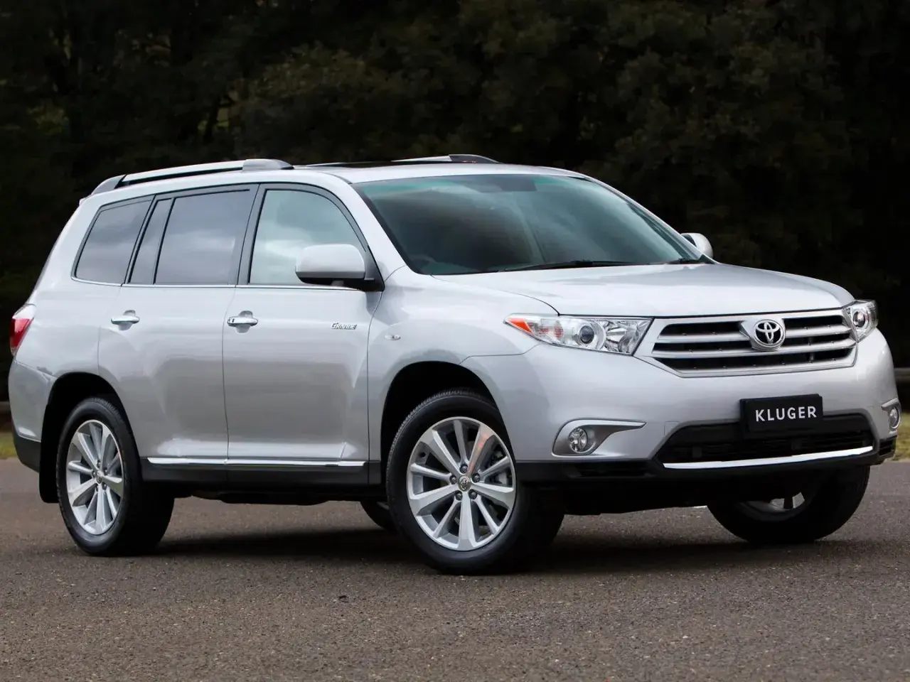 Srebrny Toyota Kluger zaparkowany na drodze, z ciemnym tłem drzew.