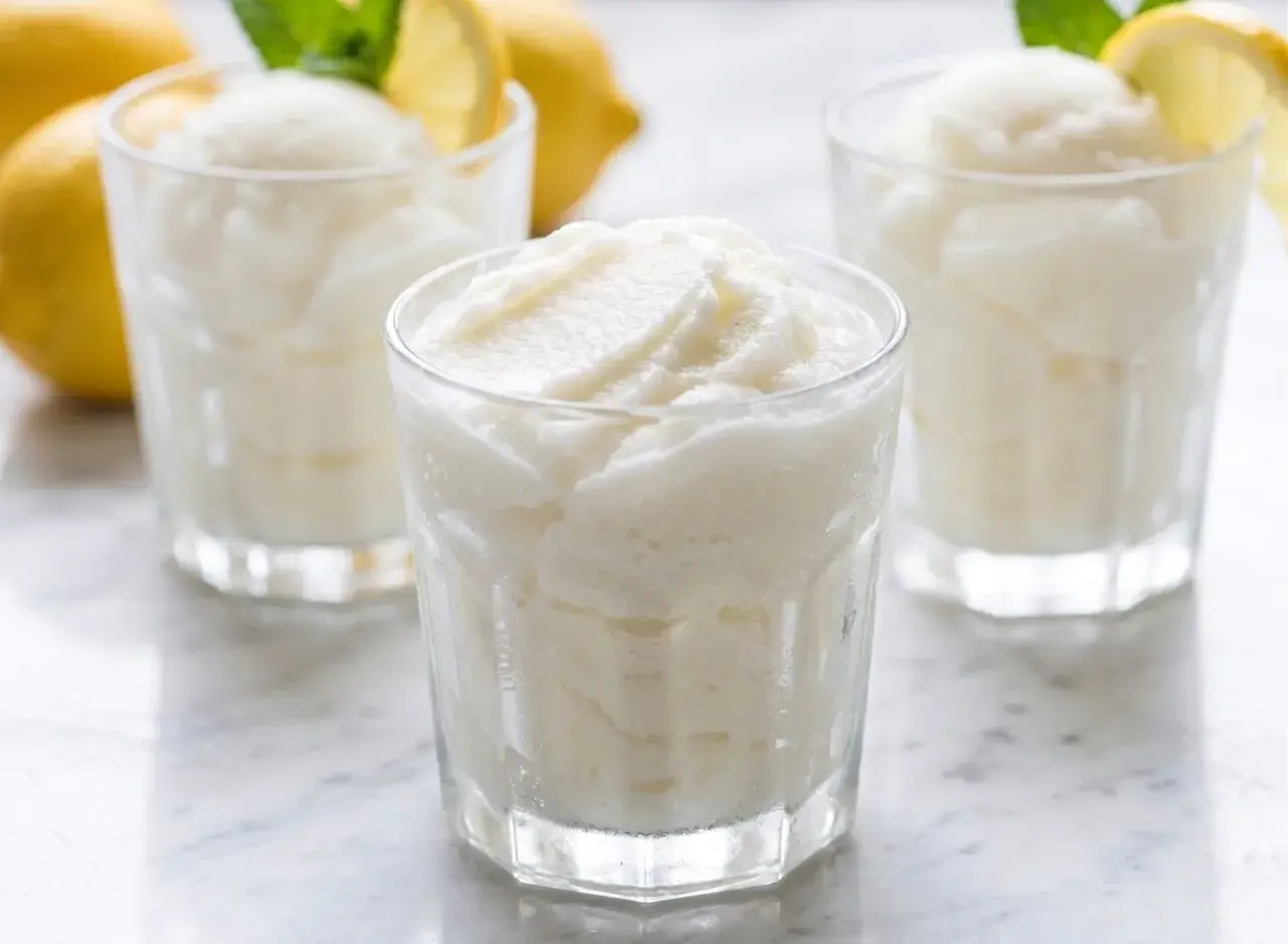 Tres vasos de sorbete de limón refrescante, con limones amarillos y hojas de menta al fondo.