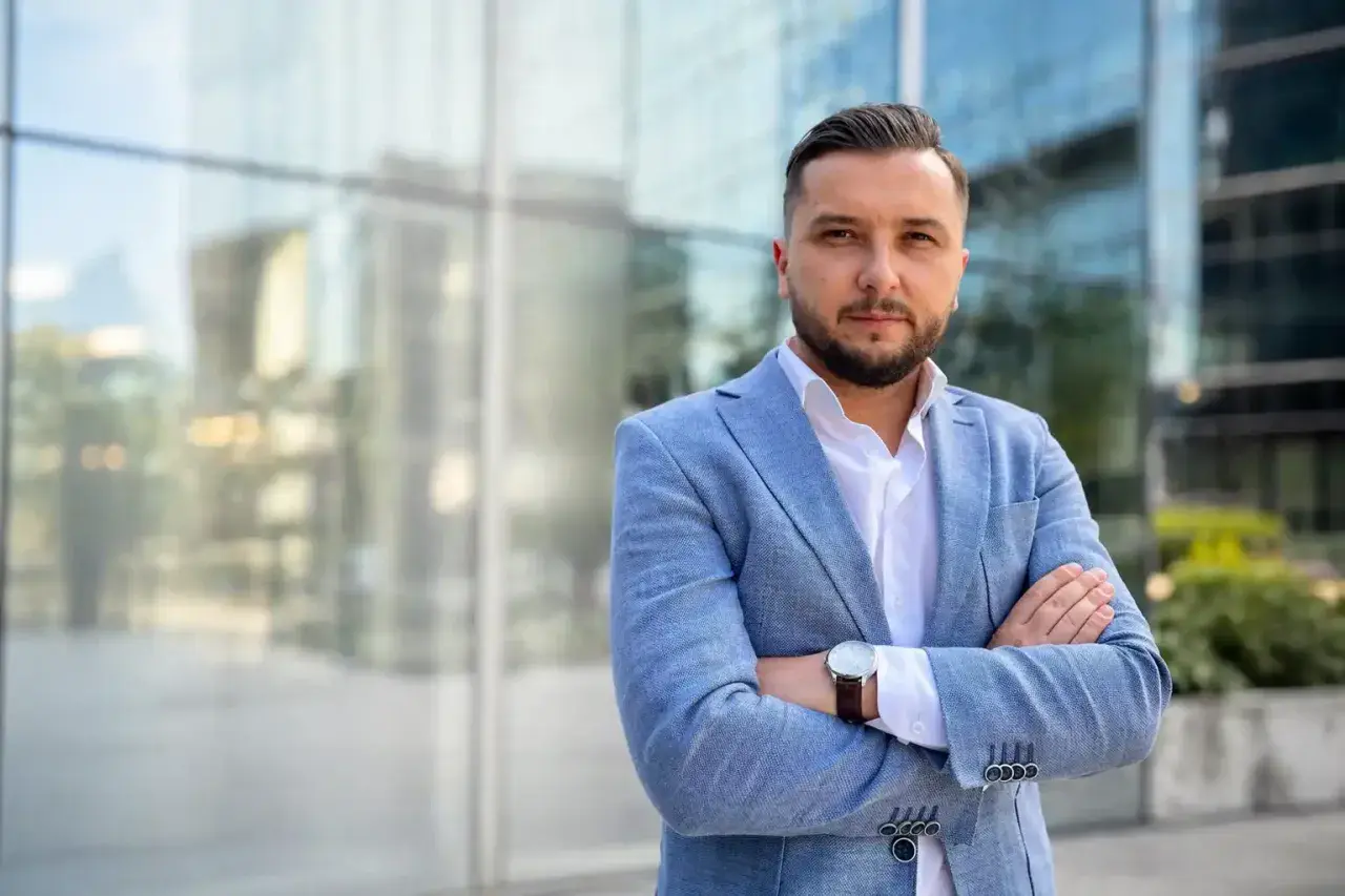 Mężczyzna w jasnoniebieskiej marynarce i białej koszuli, z rękami skrzyżowanymi na piersi, na tle nowoczesnego budynku.