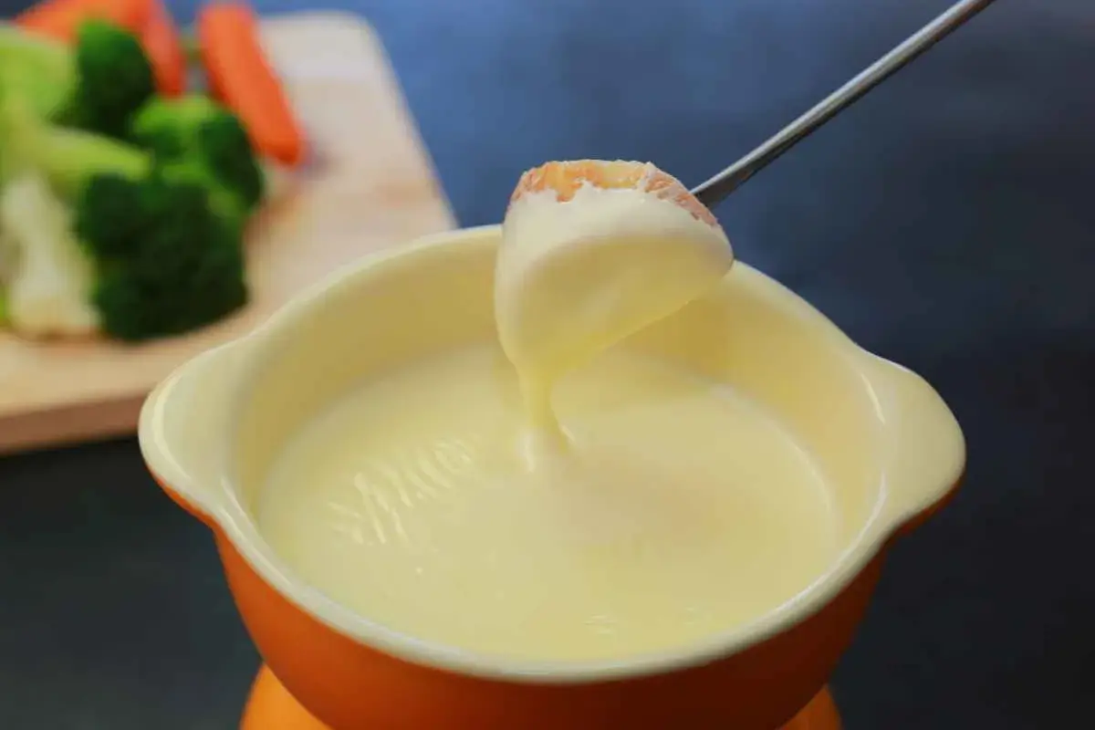 Una forchetta intinge un pezzo di pane in una cremosa fonduta per pasta. Sullo sfondo, verdure fresche.