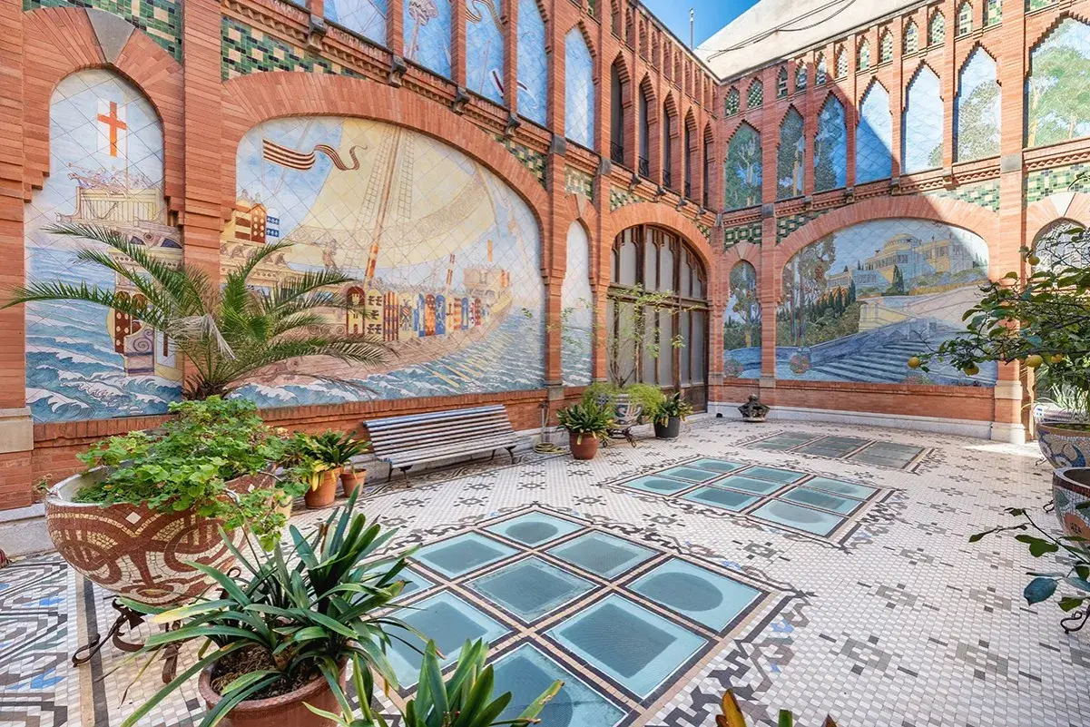 Patio de la Casa Navàs de Reus, con murales cerámicos que narran historias marítimas y paisajes, suelo de mosaico y plantas.