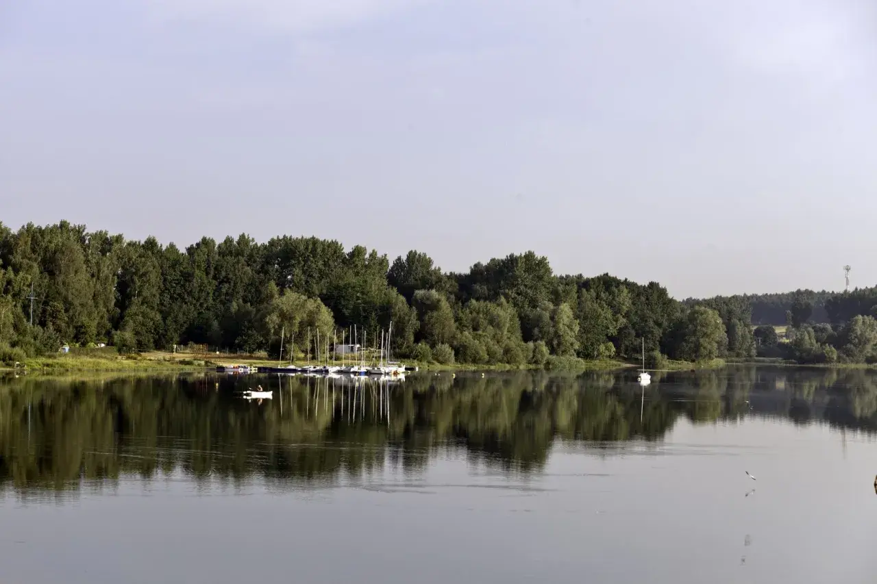 Spokojne jezioro Przeczyckie z odbijającymi się w wodzie drzewami i żaglówkami przy brzegu.