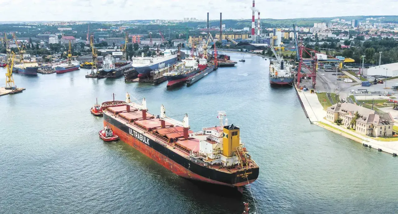 Duży statek towarowy "ULTRABULK" manewruje w porcie z pomocą holowników, w tle stocznie i miasto.