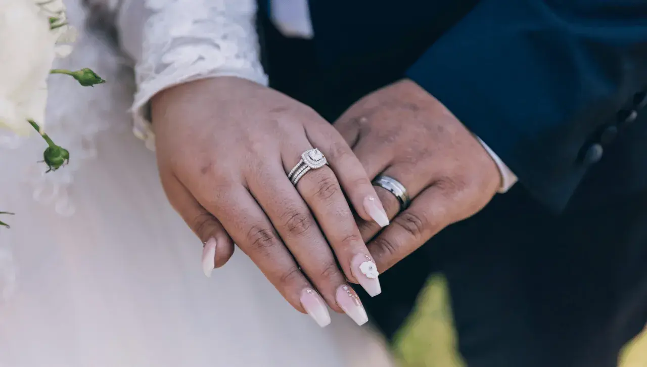 Dłonie nowożeńców z obrączkami i pięknymi paznokciami ślubnymi. Delikatny manicure zdobią kryształki i kwiat.