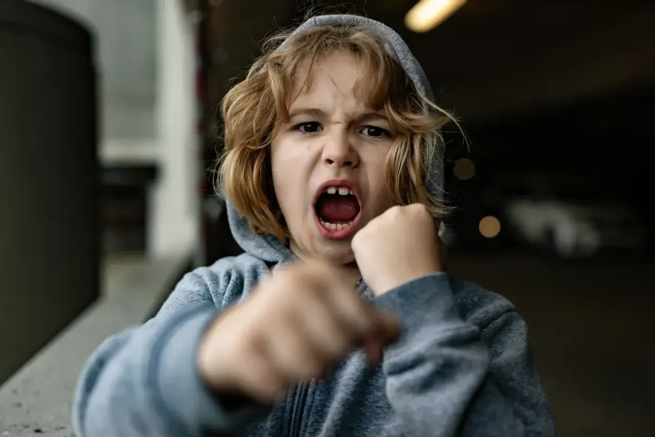 Rozgniewane dziecko w kapturze, z otwartymi ustami i zaciśniętymi pięściami, jakby chciało bić i się śmiać jednocześnie.
