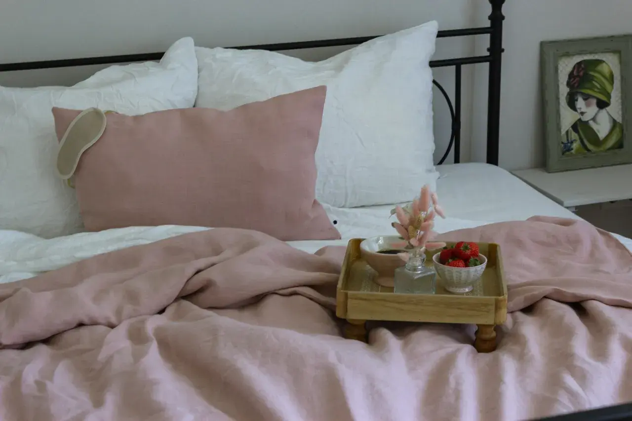Ein rosafarbenes Leinenbett mit Kissen und einer Augenmaske. Ein Tablett mit Erdbeeren und Kaffee zeigt, wie wird leinen weich.