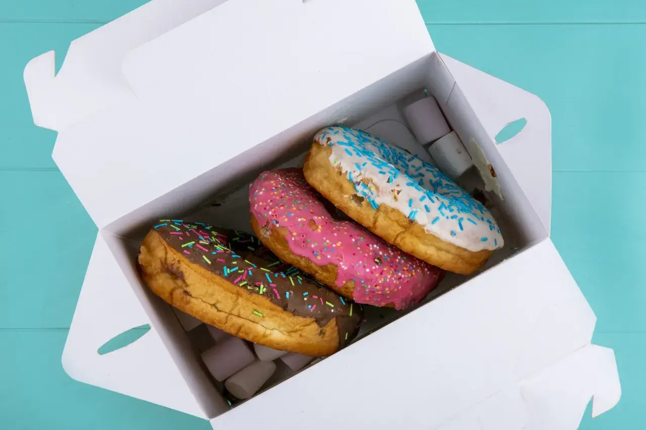 Pyszne opakowanie cukiernicze z trzema kolorowymi donutami: czekoladowym, różowym i białym z niebieską posypką.