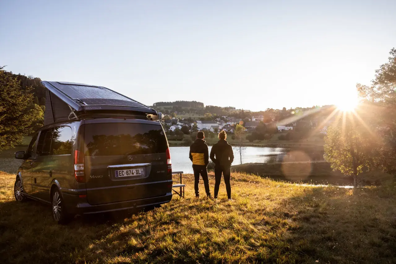 Un camping-car Westfalia avec panneaux solaires, prêt pour l'aventure près de Vulcania. Deux personnes contemplent un lac paisible au coucher du soleil.