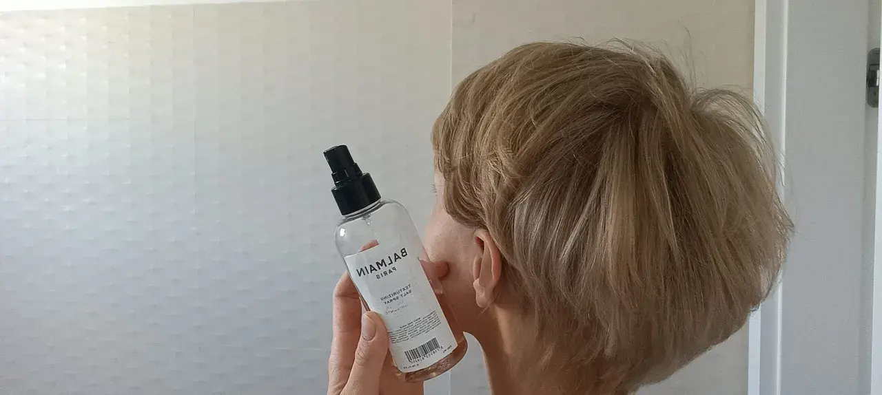 Kobieta z blond włosami trzyma butelkę z atomizerem, która zawiera sól morską do stylizacji włosów.