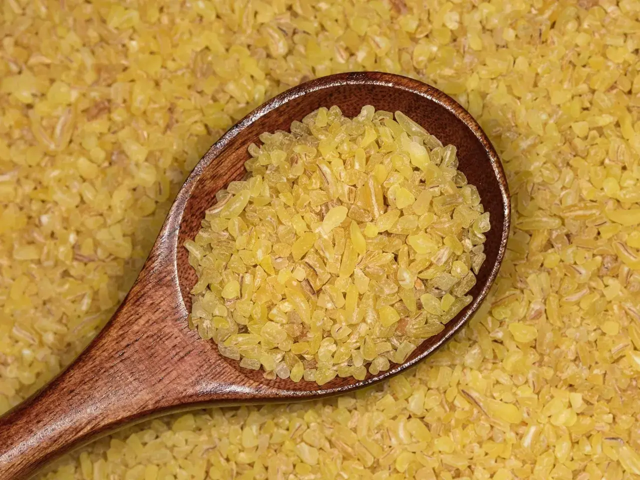 Drewniana łyżka pełna drobnej, żółtej kaszy bulgur na tle rozsypanych ziaren. To zdrowy składnik wielu dań.