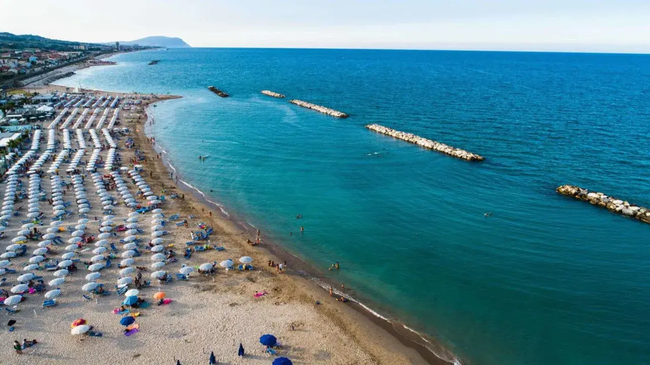 Ampia spiaggia libera a Civitanova Marche, con file ordinate di ombrelloni e lettini, bagnanti che si godono il mare turchese e dighe frangiflutti in pietra.