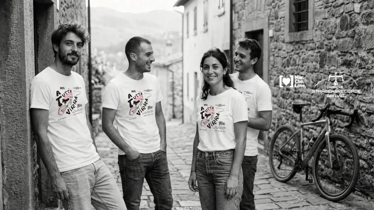 Quattro amici sorridono in un vicolo di pietra, indossando magliette con un disegno di bici e la scritta "A tutta virra". Un'espressione del vocabolario dialetto marchigiano che evoca passione.