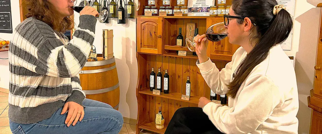 Due donne partecipano a una degustazione vini Maremma, assaporando il vino rosso tra bottiglie e prodotti tipici.