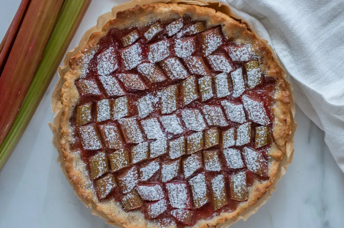 Pyszna tarta z rabarbarem, posypana cukrem pudrem, obok świeże łodygi rabarbaru.