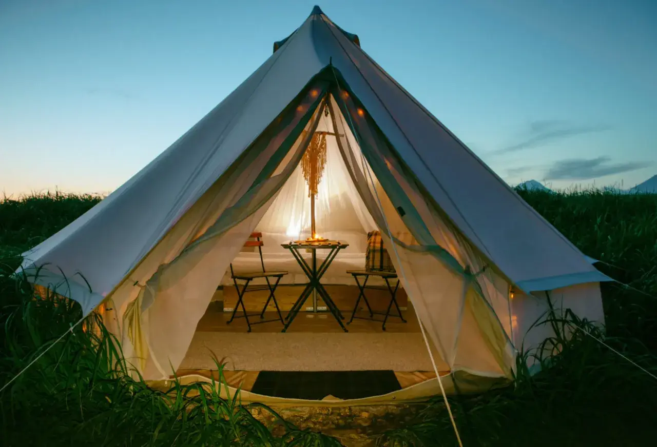 Przytulna glamping jurta oświetlona od środka, z małym stolikiem i krzesłami, otoczona zieloną trawą o zmierzchu.