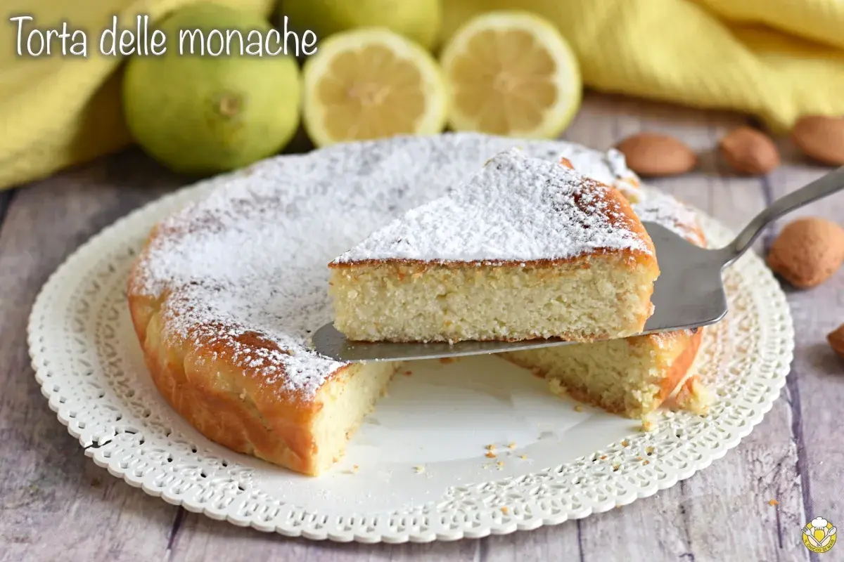 Torta delle monache soffice e profumata, cosparsa di zucchero a velo, con limoni e mandorle sullo sfondo.