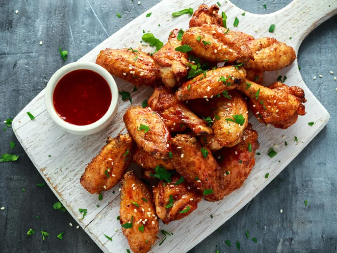 Alitas de pollo al horno jugosas, doradas y espolvoreadas con sésamo y perejil, acompañadas de una salsa roja en un cuenco.