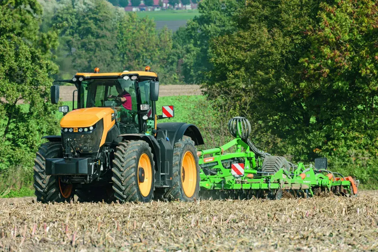 Pomarańczowy JCB Fastrac, pracując z broną, przemierza pole z prędkością 90 km/h, przygotowując ziemię do siewu.