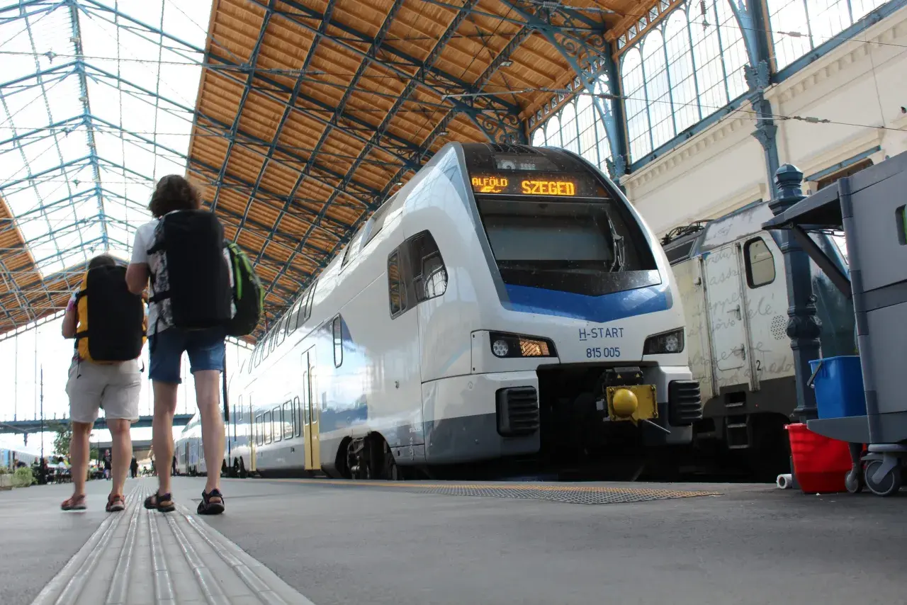 Zwei Reisende mit Rucksäcken warten auf den serbischen Zug nach Szeged unter dem beeindruckenden Dach eines Bahnhofs.