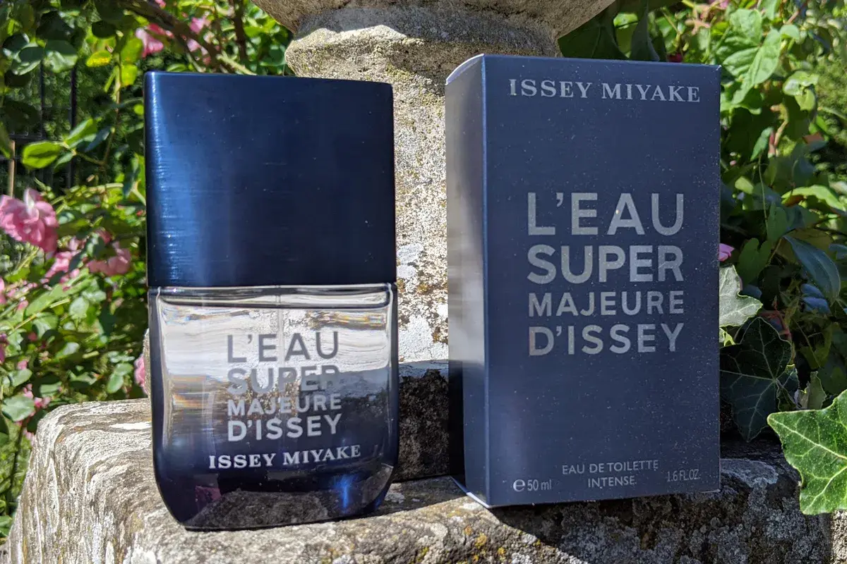 Flakon i pudełko perfum Issey Miyake L'Eau Super Majeure d'Issey na kamiennym postumencie, otoczone zielenią i różowymi kwiatami.