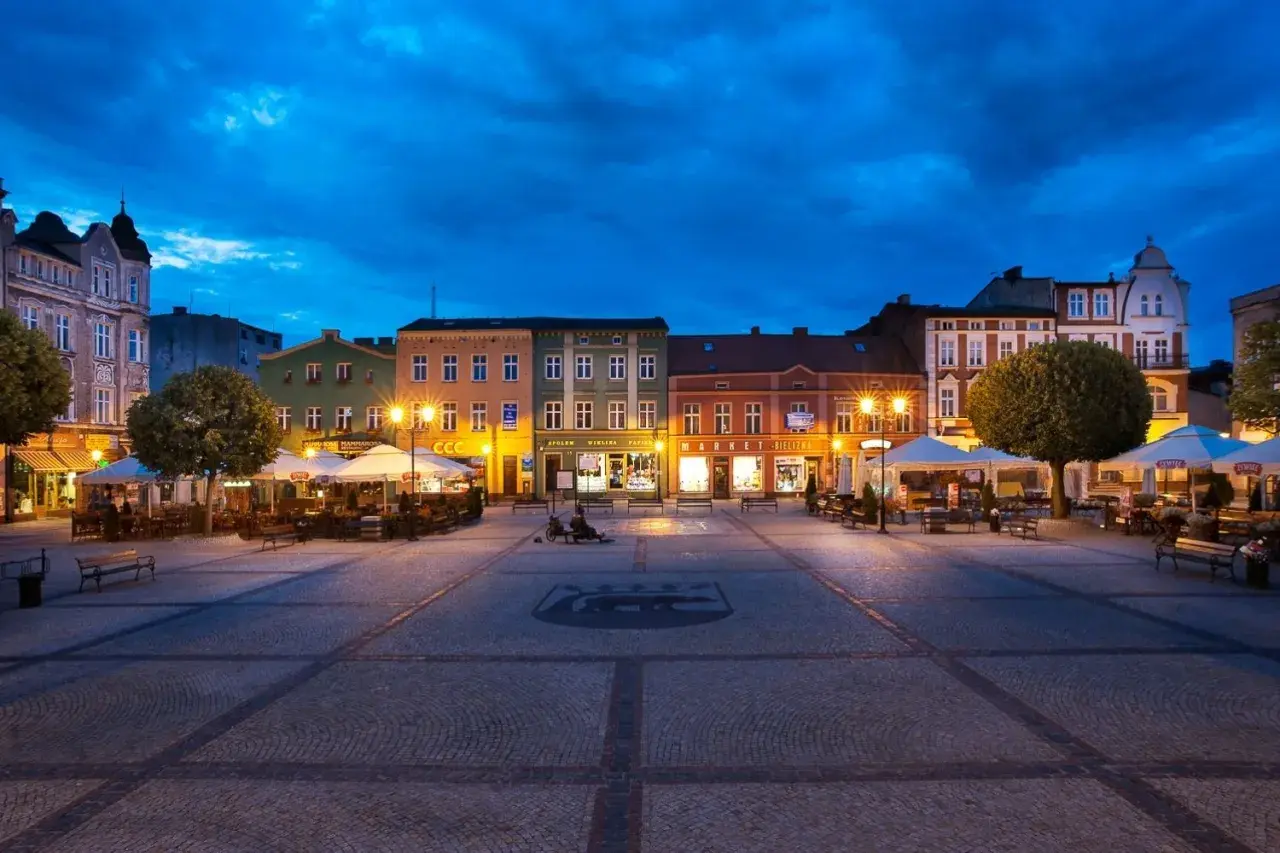 Wieczorny rynek w Kościerzynie, z oświetlonymi kamienicami i ogródkami restauracyjnymi.