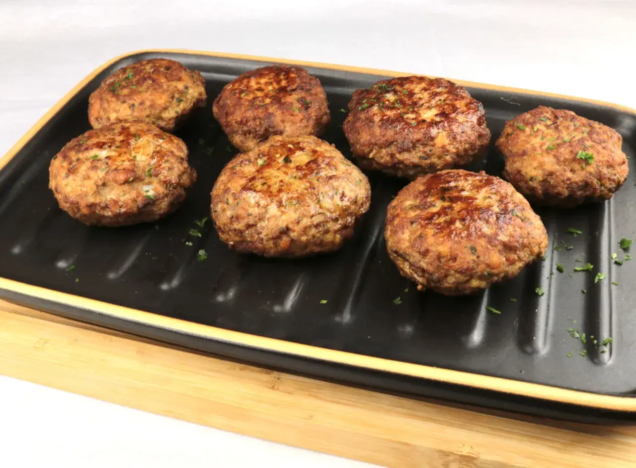 Sieben goldbraune Bouletten, wie nach Omas Rezept, liegen auf einem schwarzen Grillteller, garniert mit Petersilie.